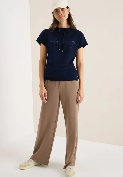 Frau mit dunkelblauem Shirt, beiger Hose und weißer Kappe steht in hellem Raum.